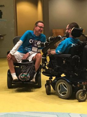 living with SMA | SMA News Today | Kevin Schaefer and Mike Blakey talk at the 2017 Cure SMA Conference. Both are seated in wheelchairs and wearing blue T-shirts.