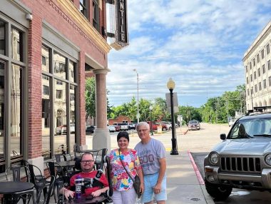 Cure SMA | SMA News Today | Kevin and his parents, Cindy and Glenn, pose for a photo outside a brick building in a downtown area of a town in Oklahoma