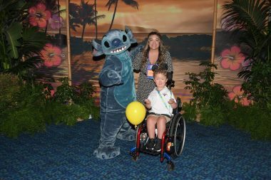 families of spinal muscular atrophy | SMA News Today | Kristen and her son Jack pose with a life-size animated figure in a costume. Jack's chair has a yellow balloon tied to it