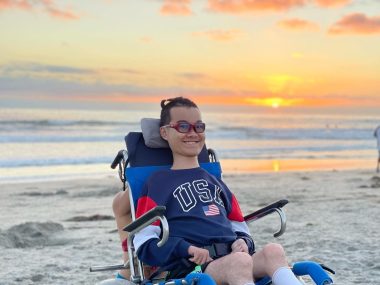 sma type 2 | SMA News Today | Ben Lou sits in his wheelchair on a beach at sunset. He's wearing a USA sweatshirt and a broad smile.