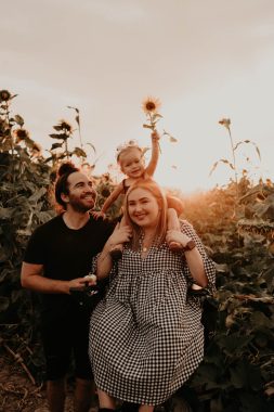 disability pride | SMA News Today | Carli Lynne Hamilton poses in a field of sunflowers with her husband, Jared, and their daughter, B. B sits on Carli's shoulders as the sun shines in the background