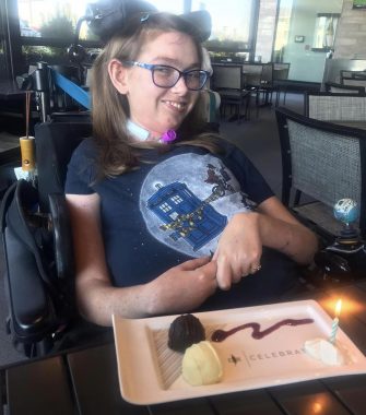 Halsey enjoys her birthday meal in a restaurant with her family. She is seated in her wheelchair, wearing a "Doctor Who" shirt, and smiling. On the table in front of her is a plate that says "Celebrate" with chocolate truffles and a candle on it.