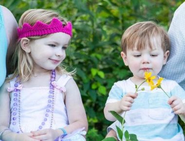 A photo of two young, blond-haired children. The girl on the left wears a purple knit crown, two necklaces of beads, and a light pink jumper. The boy wears a striped shirt in light blue and white, and he holds two flowers that look like daffodils. They appear to be outdoors and have greenery behind them. On each side are the arms of adults.
