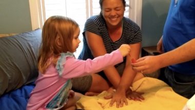 A woman and her 3-year-old niece sit cross-legged next to each other to paint fingernails. A man is just visible on the right, holding up a bottle of red nail polish. The young girl, wearing a long-sleeved pink shirt, dips the brush in the bottle. Her aunt is crossing her arms with both hands flat on the towel in front of her so the girl can paint her nails.
