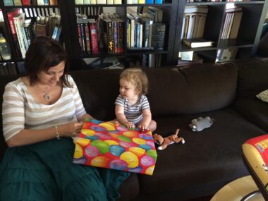A toddler sits upright next to his mother with a big, colorful birthday present in front of him and a large book case behind.