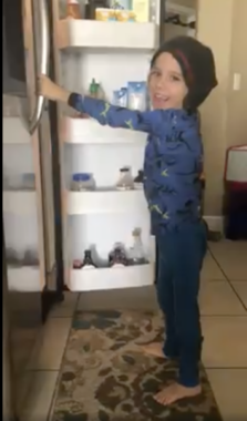 A young boy smiles as he opens a refrigerator. He is barefoot and wearing a stocking cap.