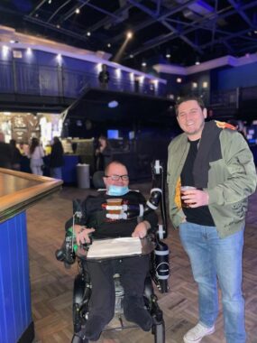 Two men, one in a wheelchair and the other standing, are inside what appears to be the lobby of a large venue. The man in the wheelchair wears glasses and is dressed in black; the standing man wears blue jeans, a black shirt, and an Army green jacket.