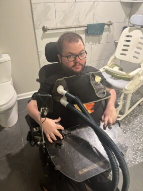 A man with close-cropped hair, glasses, a beard, and a black T-shirt is in a power wheelchair in the bathroom with a Vest Airway Clearance System on his chest for an airway clearance session to remove secretions from his lungs. The device has two large, black tubes extending from a piece that wraps around the front of his chest. He looks directly into the camera. 