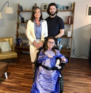A photo of a mother, son, and daughter in the living room of a home. Mom stands in back to the left, next to her son, with the daughter sitting in front. The mother has her hands folded on her daughter's wheelchair and is wearing a white coat, blue and white blouse, and white skirt; the son, a black T-shirt, which highlights his long, dark hair and black beard; the daughter is buckled into her power wheelchair and has a blue or purple long-flowing dress or blanket over her. She wears her hair long, just past the shoulders, and is wearing glasses.
