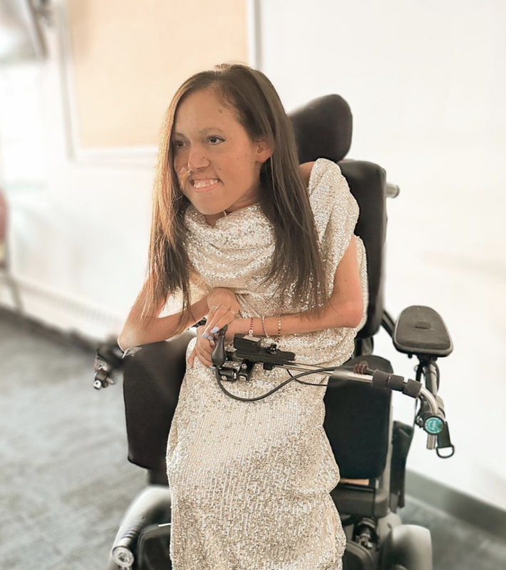 A woman with long brown hair sits in a power wheelchair. She's indoors and wearing a light gold evening gown.