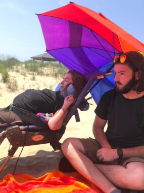 We see two people on a beach: a woman with dark hair, left, in a wheelchair, and a man with a beard, black T-shirt, and shorts, right. An orange and yellow towel is in front of him, and a multicolored umbrella is behind them both.