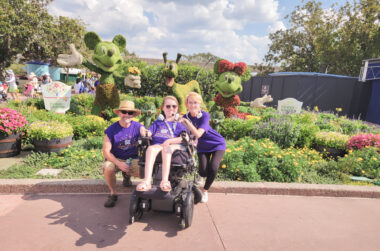 A family of three wearing purple T-shirts in support of Cure SMA pose before a Disney-themed garden at Epcot. From left to right, a father, daughter, and mother. The father wears a broad hat and sunglasses to shade from a harsh sun; the daughter wears sunglasses and is in a power wheelchair, shorts, and sandals; the mother leans on the back of the chair, with a Minnie Mouse sculpted shrub just past her left shoulder. 
