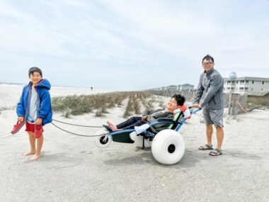 A boy reclines on a customized beach wheelchair, while his younger brother stands in front of him, pulling, and his father stands behind him, pushing.