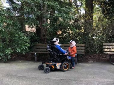 Two boys face each other next to a bench in a wooded park. Both are wearing hats and puffy jackets, and the boy on the left is in a motorized wheelchair.