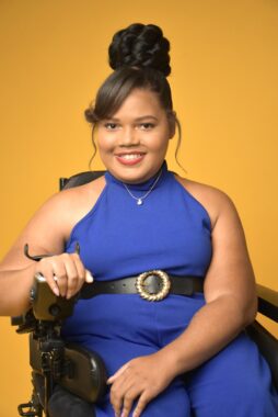 A professional photo of Candis Welch against a bright orange backdrop. She's wearing a royal blue jumpsuit with a black belt and is seated in her power wheelchair. 