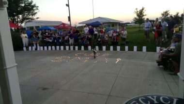 A dark photo shows people gathered in the distance with lit candles that spell "Cure SMA" on concrete. 