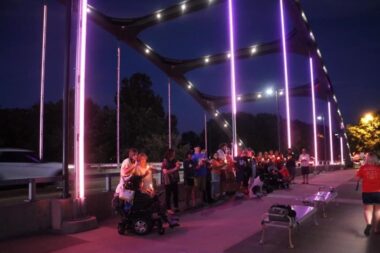 A dark and blurry photo shows people on a bridge that is lit up at night with purple and orange lights. 