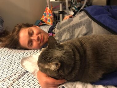 A young woman lies on her bed before a breathing treatment with her right arm outstretched. Her cat rests its head on her wrist, in the perfect position to receive some chin scratches.