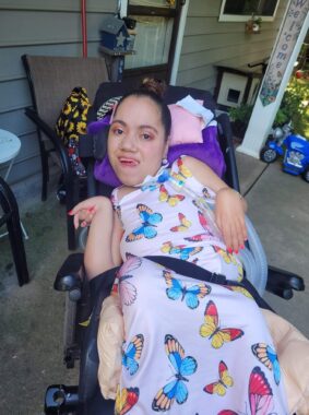 A woman wearing a pink butterfly dress smiles while sitting in her wheelchair. She appears to be outside on the patio of her home.