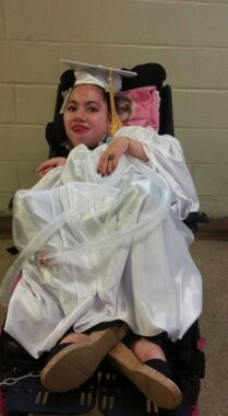 A woman in a white graduation gown and cap poses for a photo while seated in her wheelchair. 