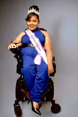 Candis Welch is crowned Ms. Wheelchair California 2023. She's wearing a royal blue jumpsuit, a tiara, and a sash, and is pictured seated in her power wheelchair.