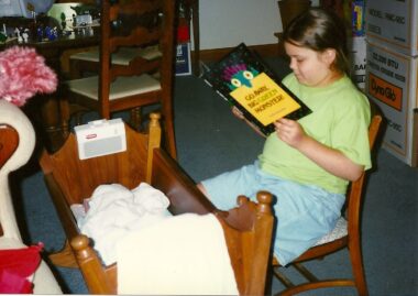 An old photo taken on film in 1997 shows a 7-year-old girl sitting on a wooden chair next to a wooden baby crib reading a child's book to a baby wrapped in blankets in the crib. 