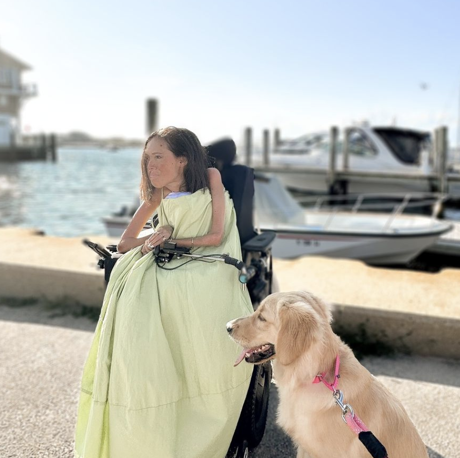 A young woman in a long green dress of pale green or dark yellow sits in a wheelchair on concrete near a dock, with boats in the background. Beside her is a smiling golden retriever, sitting and on a leash with a pink collar.
