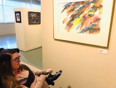 The author is pictured seated in her wheelchair in the lower left corner of the photo. She's in a well-lit art gallery and is looking at a colorful abstract painting hanging on the wall.