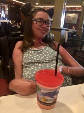 The author smiles for a photo while seated at a table in a restaurant. On the table in front of her is a plastic kids' cup, complete with a secure lid and straw for easier drinking.