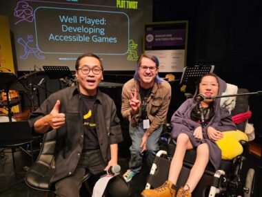 From left to right, a man dressed in black, holding a microphone; a man in a brown coat and blue jeans; and a woman in a purple hoodie. Behind them is a screen showing the words "Well Played: Developing Accessible Games."