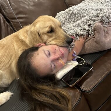 An absolutely adorable photo shows a beautiful golden retriever with a concerned look on her face lying on the couch with her head resting on the neck of a woman also lying on the couch, covered in a gray, shag blanket. The woman has a jejunostomy tube that's visible and is clearly in discomfort. Nevertheless, she is smiling due to the comfort the dog is providing her.