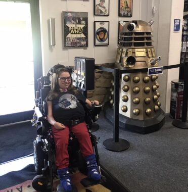 A woman peruses a "Doctor Who" retail store. She and her power wheelchair pause in front of a bunch of iconic "Doctor Who" paraphernalia. 