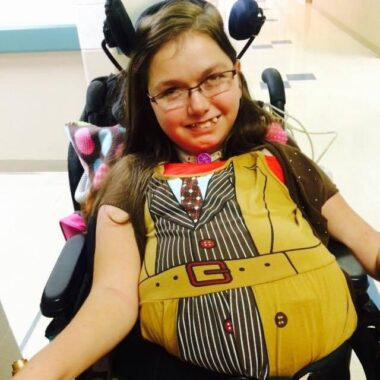 A woman wearing glasses and using a power wheelchair is dressed as a version of The Doctor from "Doctor Who." The costume is half yellow trench coat and half pin-striped suit. 