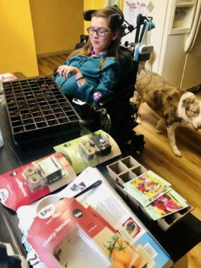 A young woman uses a plastic fork to churn small compartments of wet soil. She's seated in her power wheelchair at a table covered in soil packets and gardening supplies, and a dog is walking behind her.