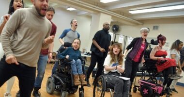 Multiple people, some using wheelchairs, sing together in a large open room.