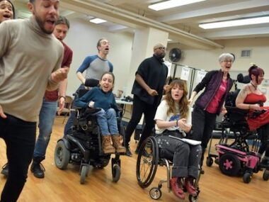 Multiple people, some using wheelchairs, sing together in a large open room.