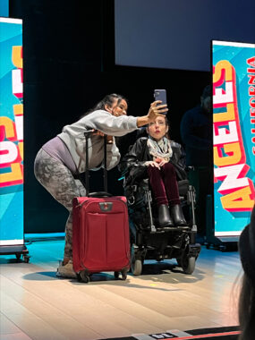 Two people on stage pose for a selfie, while one holds a suitcase.