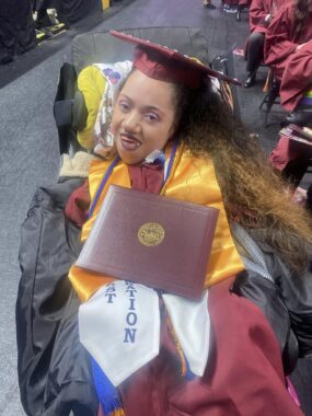 A woman is a dark red graduation cap and gown shows off her diploma. She's seated in a power wheelchair and smiling happily at the camera.