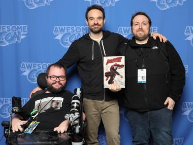 Three men are in front of a blue screen with Awesome Con logos. They're all wearing black shirts and all have beards and mustaches; the man at left is in a wheelchair, while the man at right holds up a comic book.