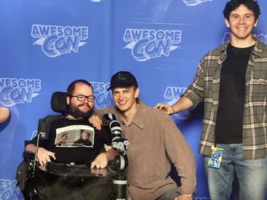 Three men in front of a blue screen featuring "Awesome Con" logos face the camera. The man at left is in a black T-shirt, is bearded and wears glasses, and is in a wheelchair; the man in the middle, in a brown shirt and navy blue baseball cap, is kneeling and has his arm on the shoulder of the seated man. At right, standing, is a young, dark-haired man in jeans, a black T-shirt, and greenish-gray flannel shirt that's worn open.