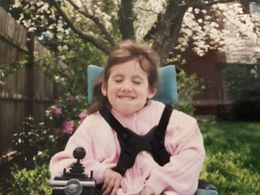 A young girl is strapped into a power wheelchair outdoors in a fenced yard, where there's green grass, low green-yellow plants, a bush with pink and white flowers, and a tree with white blooms.