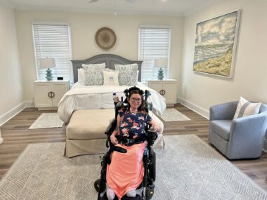 A horizontal photo with a wide frame shows a spacious bedroom with a color motif of white, tan, and light blue. In the center bottom of the photo, just in front of a bed, is a young woman in a power wheelchair with a blanket over her legs. 