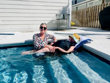 A mother assists her daughter who has SMA in a swimming pool at a rental home. The daughter lies horizontally on a mat or some type of beach chair, on the first few steps leading into the large, crystal-blue pool. It looks to be a warm, sunny day.
