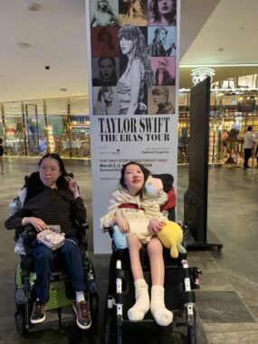 Two girls are in wheelchairs in front of a display for "Taylor Swift The Eras Tour." On the left is a young Chinese woman wearing glasses, a black and brown striped turtleneck, jeans and sneakers. Her fingers are flashing the peace sign. The young Chinese woman on the right is wearing a cream hoodie that says, “Taylor Swift: The Eras Tour” in bold, black font, which almost hides her shorts. Her smile is painted with red lipstick. 