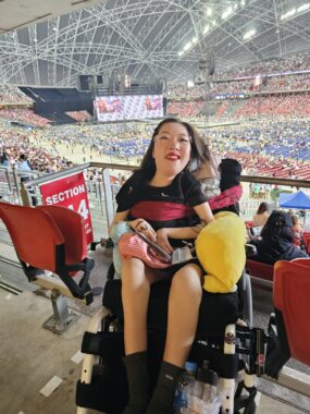 Pictured at the front of the photo is a young Chinese woman who is wearing pink glasses, red lipstick, and a black T-shirt dress with silver tassels sprinkled over it. She's in a wheelchair. Behind her is the stadium and a concert stage. 