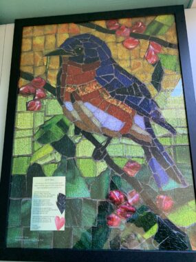 A finished jigsaw puzzle of a stained-glass piece with a bluebird sitting on the branch of a tree. In the photo, it looks like a real stained-glass piece, but it's a jigsaw puzzle. It also has some printed out lyrics, but they're too small to read in the photo. 