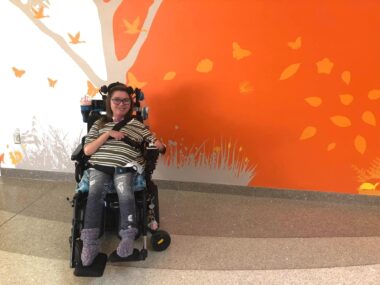 A woman in her 20s poses for a photo in front of an orange mural depicting a tree, birds, leaves, and butterflies. She's wearing a striped top and seated in her power wheelchair.