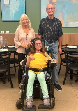 A woman in her 20s is seated in her power wheelchair, and her grandparents stand just behind her. They appear to be inside a restaurant, as tables with plates and glasses are visible in the background. All three are smiling happily.