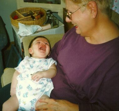 A grandmother dotes over a tiny baby she's holding in her lap. Both are smiling widely and looking at each other. The photo is from the late 1990s.