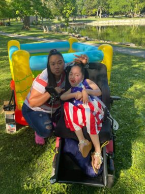 Two sisters pose outside for a photo on a sunny day. The author, Jasmine, is wearing an American flag dress and sandals and reclining in her power wheelchair. Her sister, Jessie, squats next to her, making duck lips and holding up a peace sign over Jasmine's head. There appears to be an inflatable toy right behind them, and a large pond beyond that.